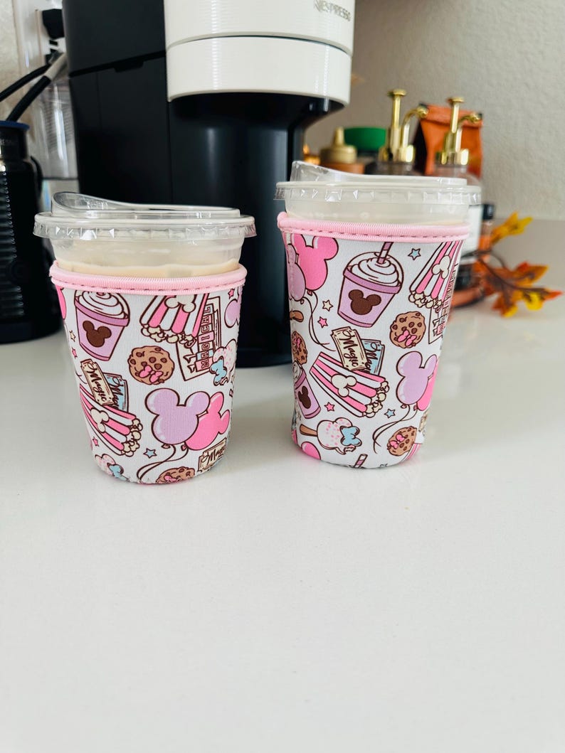 May include: Two pink and white insulated cup sleeves with a Mickey Mouse theme. The sleeves feature a variety of images including Mickey Mouse, balloons, popcorn, ice cream, and cookies. The text "Magic" is visible on one of the sleeves.
