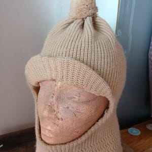 Puede incluir: Un gorro pasamontañas de punto beige con un diseño de punto acanalado y un pequeño pompón en la parte superior. El gorro cubre la cabeza, el cuello y parte de la cara. El gorro se exhibe en una cabeza de maniquí.