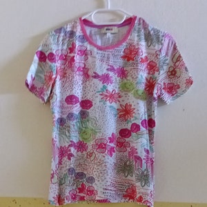 Könnte beinhalten: Weißes T-Shirt mit kurzen Ärmeln und einem rosafarbenen Blumendruck. Der Druck zeigt eine Vielzahl von Blumen in den Farben Rosa, Rot, Grün und Lila.