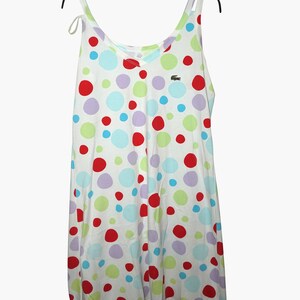 May include: White sleeveless dress with a V-neck and a polka dot pattern in red, green, blue, and purple. The dress has a small green crocodile logo on the front.
