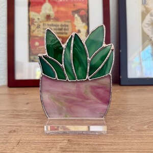 May include: A stained glass succulent plant sculpture. The plant features green leaves and a pink and white pot, all framed with silver solder. The sculpture sits on a clear acrylic base, perfect for a desk or shelf.