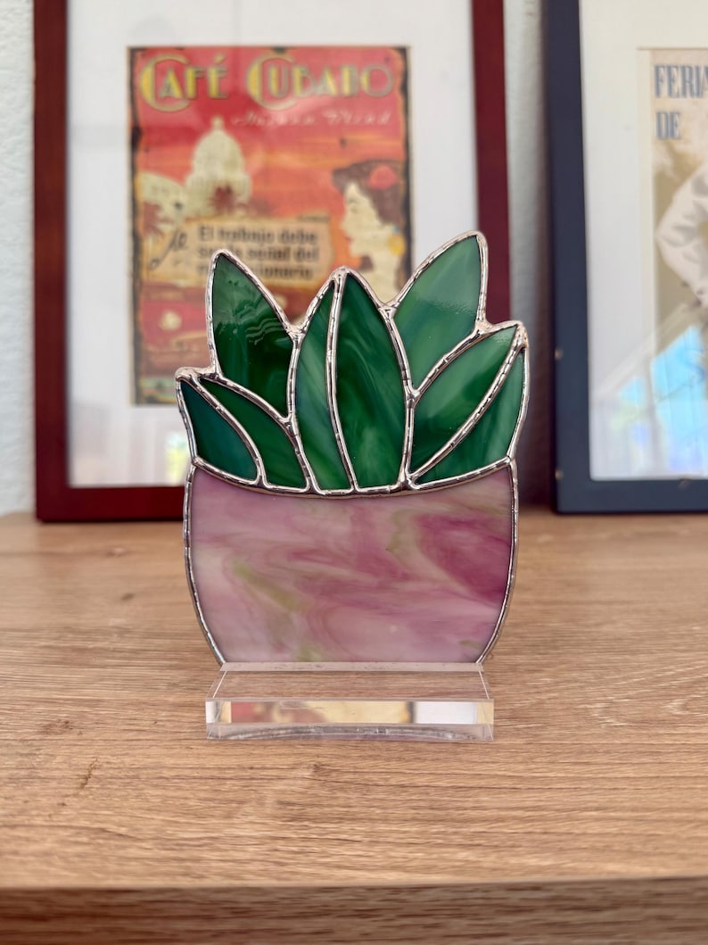 May include: A stained glass succulent plant sculpture. The plant features green leaves and a pink and white pot, all framed with silver solder. The sculpture sits on a clear acrylic base, perfect for a desk or shelf.
