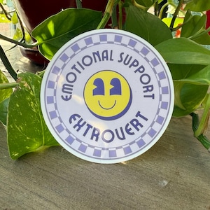 May include: A circular sticker featuring a yellow smiley face with purple eyes and a smile in the center. The text "EMOTIONAL SUPPORT" and "EXTROVERT" is printed in purple around the smiley face, surrounded by a purple and white checkerboard border.