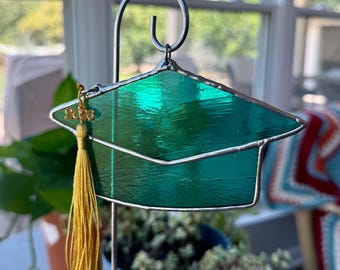 May include: A graduation cap garden stake made of green stained glass with a gold tassel. The cap is held by a silver metal frame and has a small charm with the year 2023. The stake is designed to be placed in a garden or flower pot.