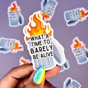 May include: A sticker featuring a cartoon trash can engulfed in flames, with the text "WHAT A TIME TO BARELY BE ALIVE" in white. The flames are orange and yellow. Several other stickers with the same design are scattered around.