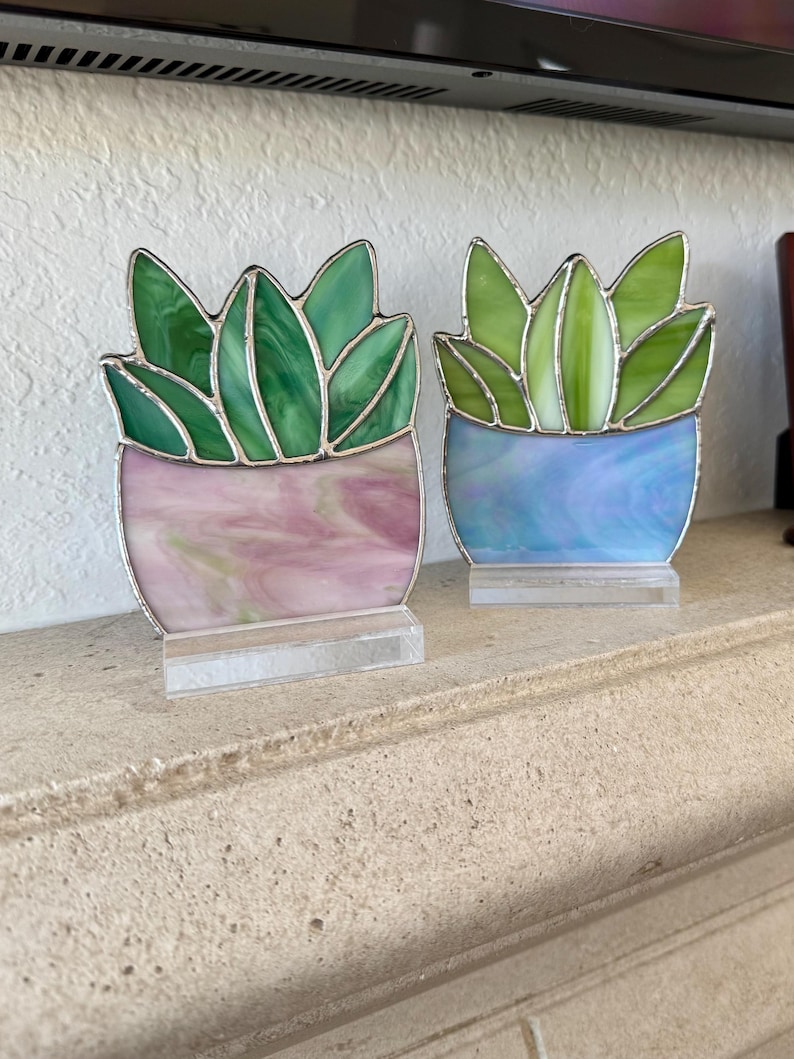 May include: Two stained glass succulent plant sculptures. One has a pink and white pot, the other a blue and white pot. The leaves are green. Each sculpture is mounted on a clear acrylic base. The plants are displayed on a stone ledge.