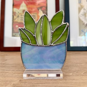 May include: A stained glass succulent sculpture. The plant has green leaves and a blue pot, set on a clear acrylic base. The glass is outlined with silver solder. The artwork is displayed on a wooden surface.