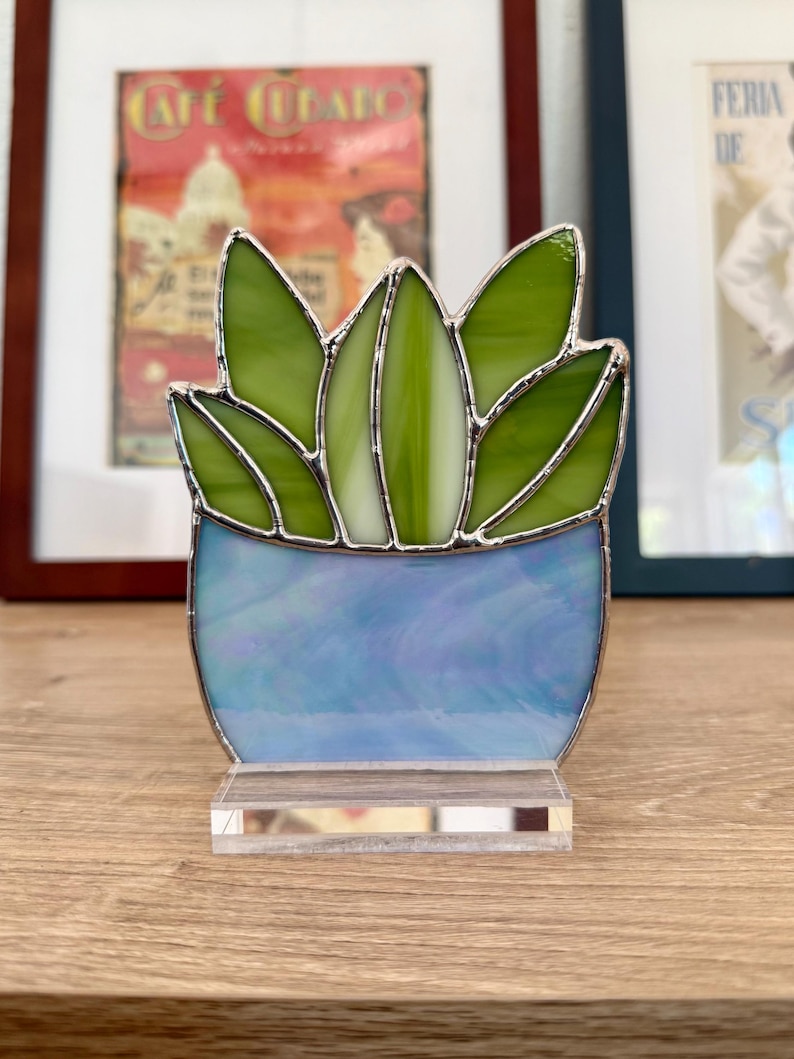 May include: A stained glass succulent sculpture. The plant has green leaves and a blue pot, set on a clear acrylic base. The glass is outlined with silver solder. The artwork is displayed on a wooden surface.