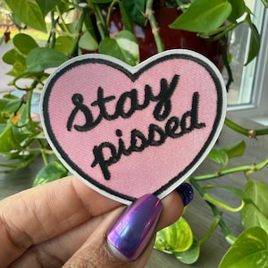 May include: A pink heart-shaped patch with the words "Stay Pissed" in black script. The patch has a white border and is held in front of a background of green plants.