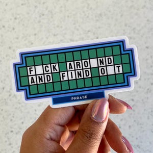 May include: A sticker with a design resembling a game show puzzle board. The text reads "F*CK AROUND AND FIND OUT" in white letters on a green background. The word "PHRASE" is written at the bottom.
