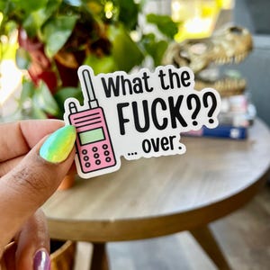 May include: A white sticker with the text "What the FUCK?? ... over." and a pink walkie-talkie illustration. The sticker is held up by a hand with colorful nail polish, against a blurred background.