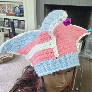 May include: A handmade crochet hat in the colors of the transgender pride flag: light blue, pink, and white. The hat has a pointed top and wide, wing-like sides. The hat is displayed on a mannequin head with dark hair.