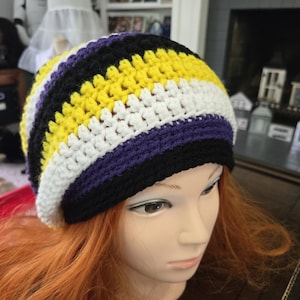 May include: A crocheted beanie featuring horizontal stripes in yellow, white, purple, and black. The hat is constructed with a textured stitch pattern and has a wide black band at the base, creating a colorful design that covers the top of the head.