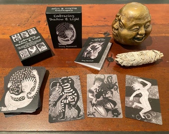 May include: A deck of black and white tarot cards with the title "Embracing Shadow & Light" on the box. The cards feature illustrations of people and animals in a stylized, graphic style. The box is on a wooden table with a white sage smudge stick and a bronze sculpture of a laughing Buddha.