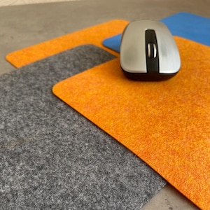 May include: Three mouse mats in different colours: grey, orange, and blue. The mouse mats are made of a soft, fuzzy material. A silver wireless mouse is sitting on top of the orange mouse mat.