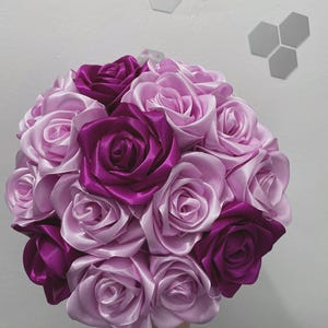 Op de afbeelding: Een boeket van kunstmatige zijden rozen in tinten paars en roze. De rozen zijn in een ronde vorm gerangschikt en worden bij elkaar gehouden met een lint.