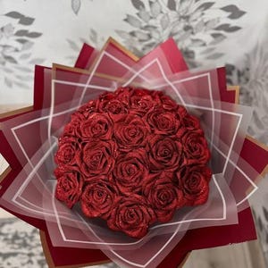 Peut inclure: Un bouquet de roses rouges pailletées, disposées en cercle. L'arrangement floral est enveloppé dans du papier rouge et blanc, avec des bordures dorées. Un cadeau élégant et coloré, idéal pour une occasion spéciale.