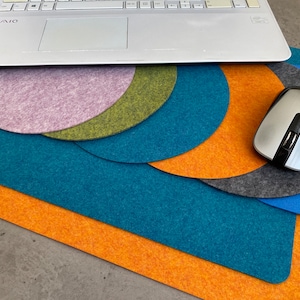 May include: A laptop sits on a collection of colourful felt desk pads. The pads are in shades of teal, orange, grey, blue, green, and lavender. A silver wireless mouse sits on top of the grey and blue pads.
