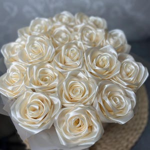 Peut inclure: Gros plan sur un bouquet de roses en satin crème. Les roses sont disposées de manière compacte en forme ronde, chaque fleur étant entièrement épanouie. Les pétales ont un éclat doux et brillant. Le bouquet est présenté dans un contenant blanc.
