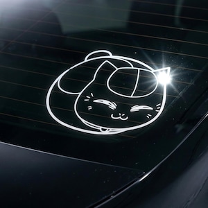 May include: A white vinyl decal sticker applied to a dark car rear window. The graphic features a stylised, circular outline of a curled-up cat with closed eyes, a small nose, and whiskers, rendered in simple, clean white lines against the glass.