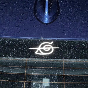 May include: White decal of a stylized leaf and swirl design, likely a car emblem, affixed to a dark surface, possibly a vehicle's rear window. The emblem is illuminated, and the background is dark blue with water droplets.