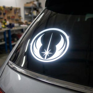 May include: A white vinyl decal of the Jedi Order symbol, a circular emblem with a central star and stylized wings, is affixed to the rear window of a silver car. The emblem is illuminated, creating a bright contrast against the dark glass.