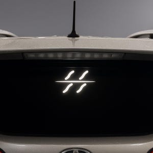 May include: A white car's rear window features a glowing, stylized symbol composed of two curved lines above and below a horizontal line. The car has a black antenna and a silver-colored body.