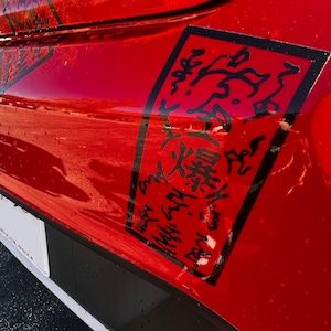May include: A close-up of a red vehicle's body with a black rectangular sticker featuring black Japanese characters. The vehicle's surface is glossy and reflects the surroundings. The sticker is on the side of the vehicle.
