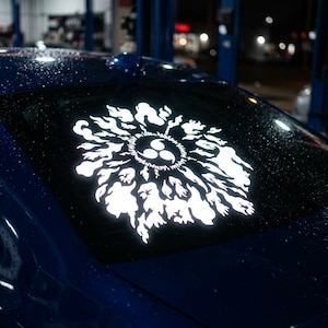 Könnte beinhalten: Weißer Vinyl-Aufkleber eines stilisierten Designs auf der Heckscheibe eines blauen Autos. Das Design zeigt ein zentrales kreisförmiges Element mit drei gebogenen Formen, umgeben von einem flammenartigen Muster. Das Auto steht nachts draußen.