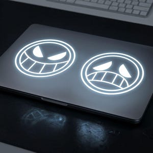 May include: A silver laptop with two glowing white circular decals. One decal features a smiling face, and the other shows a frowning face. The laptop is on a dark surface, with a keyboard visible in the background.