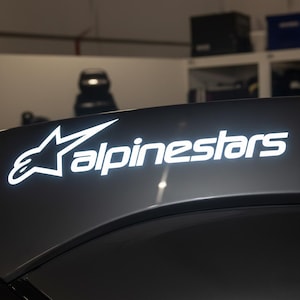 May include: Close-up of a gray surface with the white "Alpinestars" logo. The logo features a star and text, likely for motorsports apparel or gear. The image is well-lit, highlighting the brand's emblem.