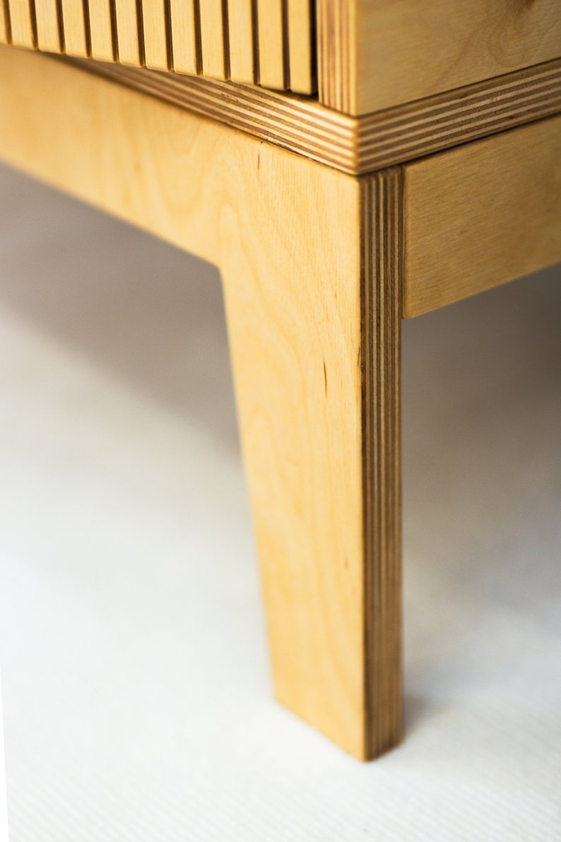 May include: Close-up of a wooden cabinet leg made of plywood. The leg is light brown and has a rectangular shape.