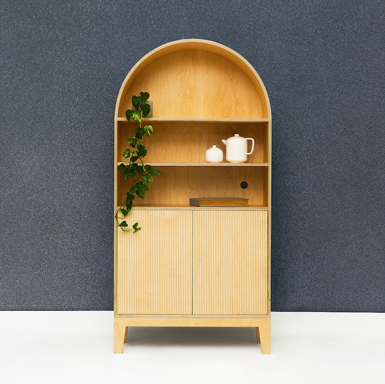 May include: A light wood arched cabinet with two doors and three shelves. The cabinet has a plant on the top shelf and two white ceramic teapots on the middle shelf.