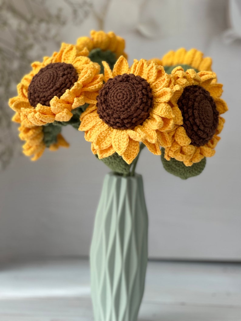 May include: A bouquet of crocheted sunflowers in a light green vase. The sunflowers are yellow with brown centers and green leaves.