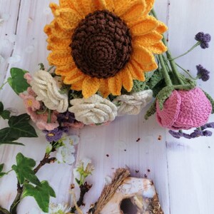 May include: A crocheted bouquet of flowers with a large yellow sunflower, white roses, pink roses, and purple flowers. The bouquet is on a white wooden background.
