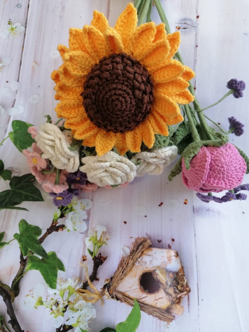 May include: A crocheted bouquet of flowers with a large yellow sunflower, white roses, pink roses, and purple flowers. The bouquet is on a white wooden background.