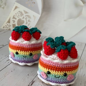 May include: Two crocheted rainbow cake toys with smiling faces and red strawberry toppings. The cakes are made with colourful yarn and have a white crocheted base.