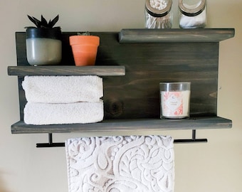 May include: A dark wood bathroom shelf with two shelves, a towel bar, and a small top shelf. The shelf has two potted succulents, a candle, and two glass jars with cotton swabs.