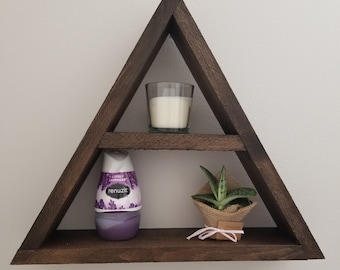 Puede incluir: Una estantería de madera triangular con tres estantes. El estante superior tiene un portavelas de vidrio transparente con una vela blanca. El estante del medio tiene una botella de ambientador púrpura con el texto "Lovely Lavender" y "Renuzit". El estante inferior tiene una pequeña planta suculenta en maceta con una envoltura de arpillera y una etiqueta rosa que dice "Gracias".