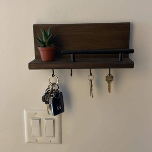 Può includere: Un porta chiavi in legno marrone scuro con una barra in metallo nero e quattro ganci. Una piccola pianta grassa in vaso si trova sullo scaffale. Un set di chiavi è appeso ai ganci.