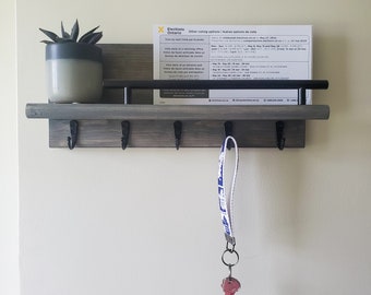 Könnte beinhalten: Ein grauer Holz-Schlüsselhalter mit vier Haken und einer schwarzen Metallstange. Ein blaues und weißes Schlüsselband mit einem roten Schlüssel hängt an einem der Haken. Eine kleine Topfpflanze mit einer Sukkulente steht auf dem Regal über den Haken. Ein weißes Papier mit Text über Wahlmöglichkeiten liegt auf dem Regal.