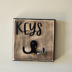 Könnte beinhalten: Ein hölzerner Schlüsselhalter mit einem Used-Look-Finish. Das Wort "KEYS" ist in schwarzer Schrift oben geschrieben. Ein schwarzer Metallhaken ist darunter angebracht. Der Schlüsselhalter ist quadratisch und hat einen dunklen Rand.