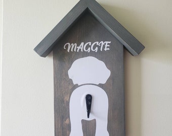 Puede incluir: Un soporte de correa para perros de madera gris con forma de casa. La palabra "MAGGIE" está escrita en blanco sobre una silueta de perro blanca. Un gancho negro está debajo.
