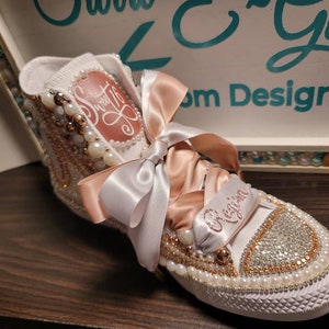 May include: White high-top sneakers decorated with pearls, rhinestones, and a pink satin ribbon. The sneakers are personalized with the name "Regina" and the words "Sweet 16".