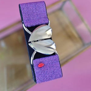 Peut inclure: Une ceinture élastique violette à paillettes avec un fermoir argenté en forme de cœur. La ceinture comporte un petit détail de lèvres rouges. La ceinture est conçue pour être portée à la taille.