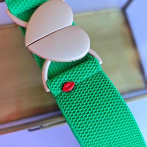 Puede incluir: Un cinturón verde brillante con una hebilla en forma de corazón de color beige claro, dividida por la mitad. Un pequeño detalle rojo en forma de labios adorna el cinturón, hecho de material tejido.