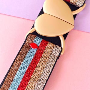 Peut inclure: Une ceinture décorative avec une boucle dorée en forme de cœur. La ceinture présente une base noire avec des rayures verticales marron, bleu, rouge et or. Un petit accent rouge est visible près de la boucle.
