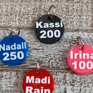 May include: Four round keychains with different names and numbers printed on them. The keychains are black, blue, red, and pink.