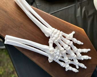 May include: A white plastic skeleton hand and forearm on a wooden surface. The hand is posed with the fingers curled.