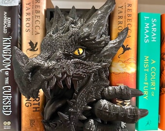 May include: A black dragon head bookend with a yellow eye, holding two books. The books are titled "Kingdom of the Cursed" and "A Court of Mist and Fury".